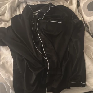 Svart satinpyjamas-skjorta med vit kant - Svart skjorta i satin med vit piping längs krage, knappslå och manschetter. Klassisk design med bröstficka och knappar framtill. Skjortan har en mjuk och glansig finish som känns lyxig mot huden. Perfekt för chill hemma eller när du vill känna dig extra comfy.