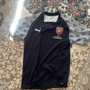 Arsenal Puma fotbollströja marinblå - Snygg Arsenal fotbollströja från Puma i marinblått med vita och svarta randiga ärmar. Tröjan har broderat Arsenal-emblem, Fly Emirates-tryck och en knapp vid halsen. Tillverkad i lätt och ventilerande polyester, perfekt för träning eller match.
