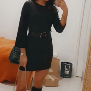 Svart kort klänning med bälte - Snygg svart kort klänning med rund hals och långa ärmar. Klänningen har ett brett bälte i midjan med metallspänne som ger en cool och markerad siluett. Perfekt för dig som gillar stilrena och enkla outfits med edge.