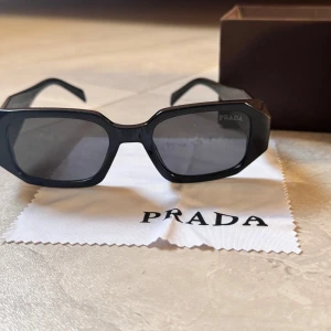 Svarta solglasögon från Prada - Snygga svarta solglasögon från Prada med rektangulära bågar och breda skalmar. Glasögonen har en cool, chunky design och kommer med originalfodral och putsduk. Perfekt accessoar för dig som vill sticka ut med en lyxig vibe.