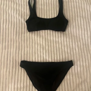 Svart ribbad bikini med breda axelband - Snygg svart bikini med ribbad struktur och breda axelband. Toppen har en fyrkantig urringning och bikinitrosan är klassisk med medelhög midja. Perfekt för strandhäng och poolparty. Materialet känns mjukt och följsamt mot huden.