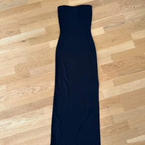 Svart lång tubklänning från NA-KD - Säljer en svart, lång tubklänning från NA-KD i storlek S. Klänningen är axelbandslös och har en slim passform som sitter snyggt mot kroppen. Materialet är mjukt och stretchigt, perfekt för dig som gillar minimalistisk stil och vill ha en enkel men stilren look.
