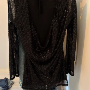 Svart glittrig långärmad klänning från design by si - Svart bklänning med glittriga paljetter som visar ryggen. Långärmad och lätt transparent med snyggt fall. Knytning i nacken för extra detalj. Perfekt för nyår eller fester