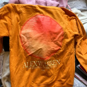  Alex Warren merch - Säljer en orange hoodie från Alex Warren med texten 'ORDINARY' på framsidan och tour-datum på baksidan. Hoodien har huva med snörning och en stor magficka. Materialet känns mjukt och är troligen bomull. Perfekt för dig som gillar färgstarka plagg och streetwear.