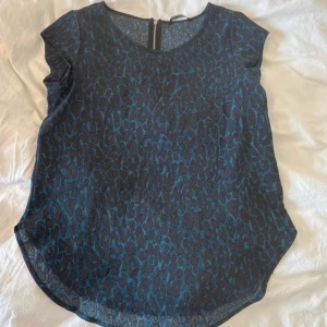 Blå leopardmönstrad blus med dragkedja - Snygg blus i blått med mörkt leopardmönster och rund hals. Toppen är ärmlös och har en silverfärgad dragkedja i ryggen. Materialet känns lätt och luftigt, perfekt för att sticka ut med ett coolt mönster. Passformen är normal och toppen är något längre baktill.