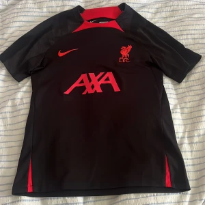 Liverpool FC svart Nike fotbollströja - Slim fit fotbollströja från Nike med Liverpool FC-logga och AXA sponsortryck i rött. Tröjan är svart med röda detaljer vid hals och sidor, tillverkad i Dri-Fit material som andas. Kortärmad och sportig design, perfekt för träning eller match. Även helt ny använd 1 gång innan 