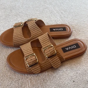 Lyxiga Beige sandaler med guldspännen från Diva - Snygga sandaler från Diva i beige med två breda remmar och guldiga spännen. Remmarna har ett diskret mönster och sandalerna har platt sula och öppen tå. Perfekt för dig som gillar stilrena och bekväma skor med lite extra detalj.