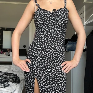 Svart blommig långklänning med slits - Supersnygg svart långklänning med vita blommor, smala axelband med volangdetaljer och en feminin slits framtill. Klänningen har figurnära passform och dragkedja i sidan för enkel påklädning. Perfekt för dig som gillar trendiga och romantiska vibes.