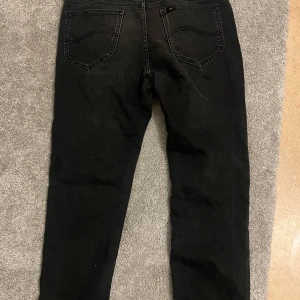 Svarta jeans från Lee - Säljer ett par svarta Lee jeans med klassisk femficksdesign och smal passform. Jeansen har en snygg mörk ton och är tillverkade i mjukt jeansmaterial med stretch för extra komfort. Perfekta för dig som gillar en clean och stilren look.