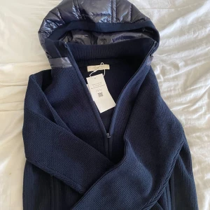 Mörkblå stickad jacka från Syless - Snygg mörkblå stickad jacka från Syless i storlek XXS. Jackan har en avtagbar glansig huva i syntetmaterial och två dragkedjeförsedda fickor framtill. Perfekt för dig som gillar stilrena detaljer och vill ha något som sticker ut lite extra.