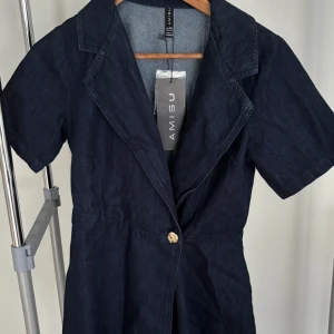 Mörkblå jeansklänning från Amisu - Snygg mörkblå jeansklänning från Amisu med korta ärmar och klassisk krage. Klänningen har en guldig knapp framtill och är tillverkad i jeansmaterial som ger en cool och tidlös vibe. Perfekt för dig som gillar denim och vill ha en enkel men stilren look.