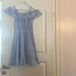 Blåvit randig kortklänning H&M - Supersöt blåvit randig klänning från H&M med volangkant och smala axelband. Offshoulder-stil med dekorativa små bollar längs volangen. Perfekt för varma dagar och har en luftig passform med resår i midjan. Materialet är mjuk bomull.