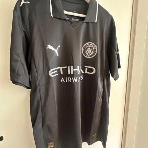 Manchester City Bortaställ 25/26 Fin Kvalité - Snygg svart Manchester City fotbollströja från Puma med silverfärgade detaljer och klubbmärke på bröstet. Tröjan har korta ärmar, klassisk krage med vit kant och är tillverkad i lätt, ventilerande polyester. Etihad Airways-logga framtill och autenticitetsmärke nedtill.