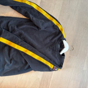 Svart fleece-tröja Parajumpers gul detalj - Svart fleece-tröja från Parajumpers i storlek L i barn storlek så ungefär xs med coola gula ränder längs ärmarna och broderad patch på axeln. Tröjan har ribbade muddar och orange söm vid nederkanten. Perfekt för dig som gillar sportig och street-inspirerad stil.