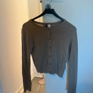 Brun kofta från H&M med knappar - Säljer en enkel och stilren brun kofta från H&M. Koftan har långa ärmar, rund hals och knäppning med små knappar framtill. Materialet känns mjukt och lätt, perfekt att slänga på sig över en t-shirt eller linne.