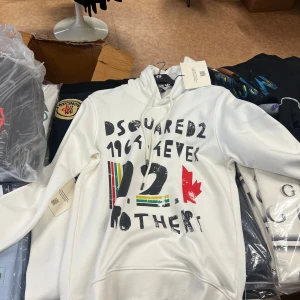 Vit hoodie från Dsquared2  - Säljer en vit hoodie från Dsquared2 med coolt grafiskt tryck i svart, rött och flera färger på framsidan. Tröjan har huva med snörning och är tillverkad i 100% bomull. Perfekt för dig som gillar streetwear och vill sticka ut med en snygg design.