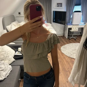 Zara topp - Säljer en beige croppad topp med omlott och volangdetaljer. Toppen har en oneshoulder-design med en axel bar och den andra täckt av volang. Materialet är mjukt och lätt, perfekt för varma dagar. Passar dig som gillar trendiga och feminina plagg. Lappen är kvar