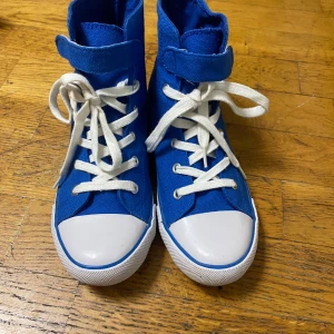 Blå höga sneakers med vit tå - Snygga blå höga sneakers med vit rund tå och platt sula. Skorna har vita snören och en bred kardborre upptill för extra stilpoäng. Tillverkade i canvasmaterial som ger en cool och avslappnad vibe. Perfekta för dig som gillar streetstyle och vill sticka ut.