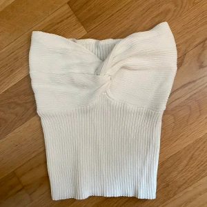 Vit ribbad bandeau tubtopp - Säljer en vit bandeau tubtopp med snygg twistad detalj framtill. Toppen är ribbad och har en tight passform som sitter skönt mot kroppen och väldigt stretchig. Perfekt att styla med jeans eller kjol för en clean look.