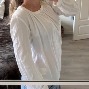 Vit blus med rynkade detaljer - Säljer en vit blus med rynkade detaljer vid halsringningen och långa, luftiga ärmar. Blusen har en avslappnad passform och är gjord i ett tunt, mjukt material som känns skönt mot huden. Perfekt att matcha med jeans för en fräsch look💕