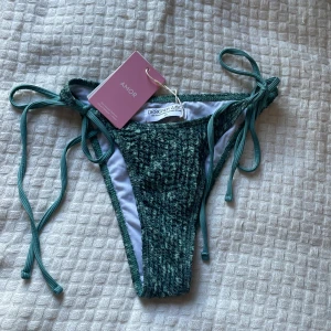 Grön bikiniunderdel från Design By Amor - Supersnygg grön bikiniunderdel från Design By Amor med ribbad struktur och knytband i sidorna. Materialet är mjukt och stretchigt syntet, perfekt för strandhäng. Färgen är djup grön med en cool textur som ger extra vibe. Det finns en överdel till så skriv om ni vill se den också! Bara underdelen kostar 50 kr och med överdelen blir det 90 kr! 