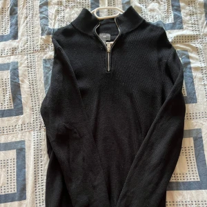 Svart stickad tröja med half zip H&M - Svart stickad tröja från H&M med ribbad struktur och half zip-dragkedja vid halsen. Tröjan har långärmad design och en hög krage som ger en clean och stilren look. Perfekt för dig som gillar enkla och snygga plagg.