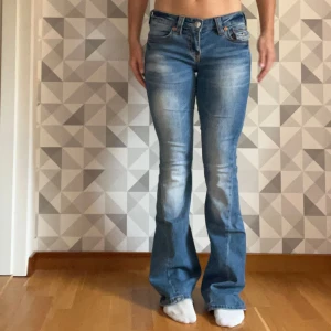 True Religion Joey Low Rise Flare jeans - Säljer ett par blå Joey Low Rise Flare jeans från True Religion. De har låg midja, bootcut och snygga detaljer på bakfickorna med kontrastsömmar. Jeansen är i klassisk denim med slitningar framtill och bak, perfekt för dig som gillar retrovibe och statement-plagg. Använt fåtal gånger. Storlek 25. Nypris 1600