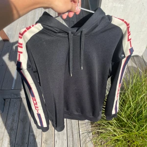 Svart gucci hoddie  - Svart hoodie från Gucci med huva och snörning. På ärmarna finns breda vita band med röd GUCCI-logga och blå detaljer. Tröjan har lång ärm och en loose passform, perfekt för dig som gillar streetwear och exklusiva märken. Materialet känns mjukt och skönt mot huden.