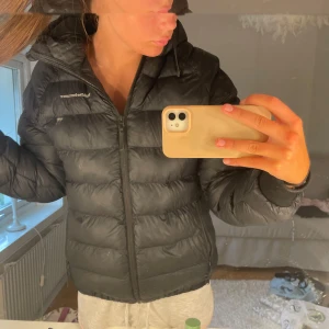 Svart dunjacka från Peak Performance - Säljer en svart dunjacka från Peak Performance med huva och dragkedja framtill. Jackan har en puffad look, långärmade ärmar och diskret logga på bröstet. Perfekt för kyliga dagar och enkel att matcha med olika outfits.