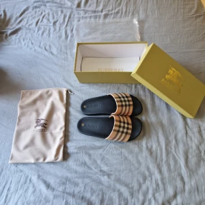 Burberry sandaler rutig beige/svart - Säljer ett par stilrena Burberry sandaler i storlek 43. De har klassiskt rutigt mönster i beige, svart och brunt på remmen och en svart platt sula med Burberry-logga. Perfekta för sommaren och riktigt snygga till shorts eller jeans.