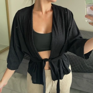 Svart omlott overshirt med knytband - Svart omlott overshirt från Lindex med knytband i midjan och diskret glittrig finish. 