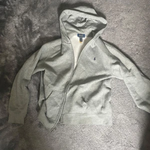 Grå hoodie från Polo Ralph Lauren  - Säljer en grå hoodie från Polo Ralph Lauren i storlek XS (XL barnstorlek). Tröjan har huva, dragkedja framtill och två fickor. Materialet är mjukt och skönt, perfekt för chill dagar. Klassisk design med liten Polo-logga på bröstet och ribbade muddar vid ärmslut och nederkant.