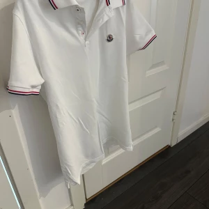 ÄKTA Vit Moncler pikétröja med röd/blå kant - Använd 3-4 GÅNGER ENDAST. Storlek S men rekommendert är XS. ÄKTA Vit pikétröja från Moncler med klassisk krage och korta ärmar. Tröjan har Moncler-logga på bröstet och snygga rödblå ränder på krage och ärmslut. Tillverkad i bomull för skön känsla och stilren look. Perfekt för dig som gillar exklusiva märken.