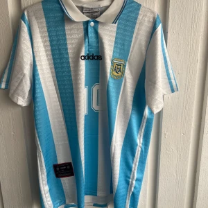 Argentina Maradona 10 Adidas fotbollströja - Säljer en klassisk Argentina fotbollströja från Adidas med Maradona och nummer 10 på ryggen. Tröjan har blå och vita vertikala ränder, korta ärmar och krage med knapp. Materialet är lätt polyester, perfekt för match eller samling.