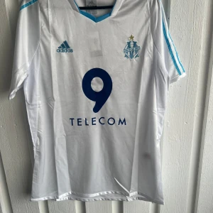 Marseille Drogba fotbollströja Adidas - Snygg vit Marseille fotbollströja från Adidas med blå detaljer och tryck. Framsidan har klubbmärke, Adidas-logga och nummer 9, medan baksidan har DROGBA och nummer 11 i blått. Klassisk V-ringning och korta ärmar. Perfekt för fans av Drogba och OM.