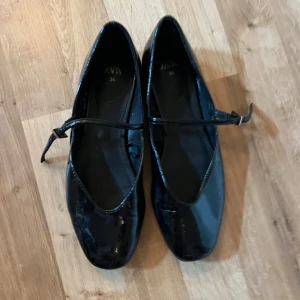 Svarta ballerinaskor från Zara - Snygga svarta ballerinaskor från Zara med rund tå och en elegant rem över foten som stängs med ett litet spänne i guld. Skorna har en glansig finish och är tillverkade i syntetmaterial som ger en cool look. Perfekta för dig som gillar stilrena och klassiska skor.