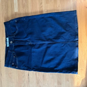 Mörkblå jeanskjol Miss Natalie - Snygg mörkblå jeanskjol från Miss Natalie med klassisk femficksdesign och kontrastsömmar. Kjolen har knapp och dragkedja framtill samt bälteshällor. Perfekt för dig som gillar enkel och tidlös denimstil.