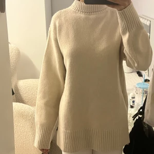 Beige stickad polotröja med slits - Säljer en beige stickad polotröja med ribbad hög krage som har en snygg slits framtill. Tröjan har långa ärmar och ribbade muddar vid ärmslut och nederkant. Passformen är loose och perfekt för lager-på-lager under höst och vinter.