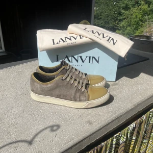 Lanvin sneakers i mocka och lack - Lanvin sneakers med grå mocka och olivgröna lackade tår. Klassisk låg modell med rund tå, snörning och vit platt sula. Snygga kontraster mellan material och färg, perfekt för dig som gillar exklusiva detaljer och fransk design.