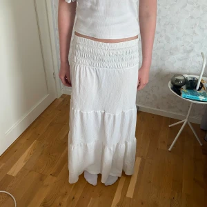 Vit långkjol från Gina Tricot - Supersöt vit långkjol från Gina Tricot med smockad midja och volanger. Kjolen är gjord i ett lätt och luftigt tyg, perfekt för varma dagar. Den har en flowy passform och är riktigt skön att bära. Passar dig som gillar en enkel men stilren look.