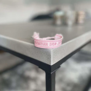 Rosa Christian Dior J'Adior armband - Säljer ett snyggt rosa armband från Christian Dior med texten 'J'Adior' broderad i ljusrosa. Armbandet är gjort i ett mjukt tygmaterial och har justerbar knytning med tofsar i ändarna. Perfekt för att ge din outfit en lyxig och trendig touch.