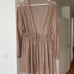 Champagnefärgad glittrig strandklänning  - Säljer en superfin strandklänning i champagnefärg med glittriga ränder. Klänningen har knyt i midjan. Materialet är lätt och skimrande