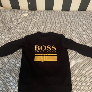 Svart sweatshirt från Hugo Boss - Svart sweatshirt från Hugo Boss med stor guldfärgad BOSS-logga och två guldfärgade linjer på bröstet. Tröjan har rund hals, långa ärmar och ribbade muddar. Perfekt för dig som gillar stilrena och lyxiga streetwear-plagg. Det är en storlek S och aldrig varit använd. 