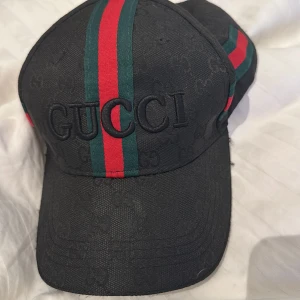 Svart Gucci keps med röd/grön rand - Snygg svart keps från Gucci med klassiska broderade loggan framtill och ikoniska röd/gröna ränder som går över hela kepsen. Justerbar rem baktill med metallspänne. Materialet är bomull och kepsen har diskret mönster med GG-loggor över hela ytan.