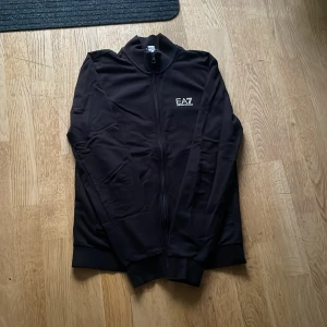 Svart track jacket från EA7 Emporio Armani - Säljer en svart track jacket från EA7 Emporio Armani med dragkedja framtill och logga på bröstet. Jackan har hög krage och ribbade muddar vid ärmslut och nederkant. Perfekt för en sportig och stilren look. Den är använd max 3 gånger och har legat i garderoben ett tag så den är skrynklig, men det går att fixa! Priset kan diskuteras. (Köpt på Boozt) STORLEK S