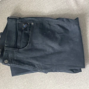 Svarta jeansbyxor från Replay - Säljer ett par svarta jeansbyxor från Replay med klassisk femficksdesign och knappgylf. Byxorna har en rak passform och är perfekta för en stilren look. Diskret Replay-logga vid fickan.