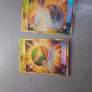 Pokémonkort: Switch och Nest Ball - Två Pokémonkort med motiv av Switch och Nest Ball. Båda korten har en glittrig, holografisk yta med starka färger som gult, orange och grönt. Perfekt för samlare eller Pokémon-entusiaster som vill utöka sin kortsamling.