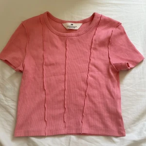 Rosa ribbad t-shirt från H&M - Säljer en söt rosa ribbad t-shirt från H&M med korta ärmar och rund halsringning. Toppen har dekorativa sömmar framtill som ger en cool touch. Perfekt till jeans eller kjol!