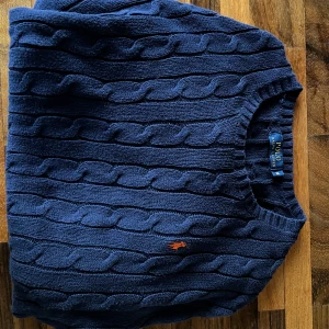 Mörkblå tröja från Polo Ralph Lauren - Säljer en klassisk mörkblå kabelstickad tröja från Polo Ralph Lauren med rund halsringning och broderad logga i orange på bröstet. Tröjan är i bomull och har långa ärmar. Perfekt för lager-på-lager-stil.