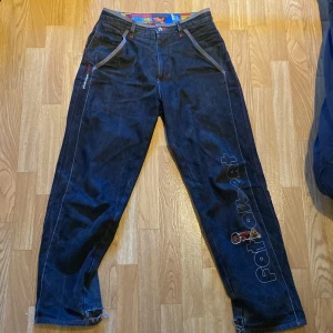 Mörkblå jeans från Platinum Fubu med broderad text - Säljer ett par mörkblå jeans från Platinum Fubu med broderad text längs ena benet och färgglatt innerfoder. Jeansen har raka ben, klassiska fickor och råa, fransiga benslut för en streetwear-look.